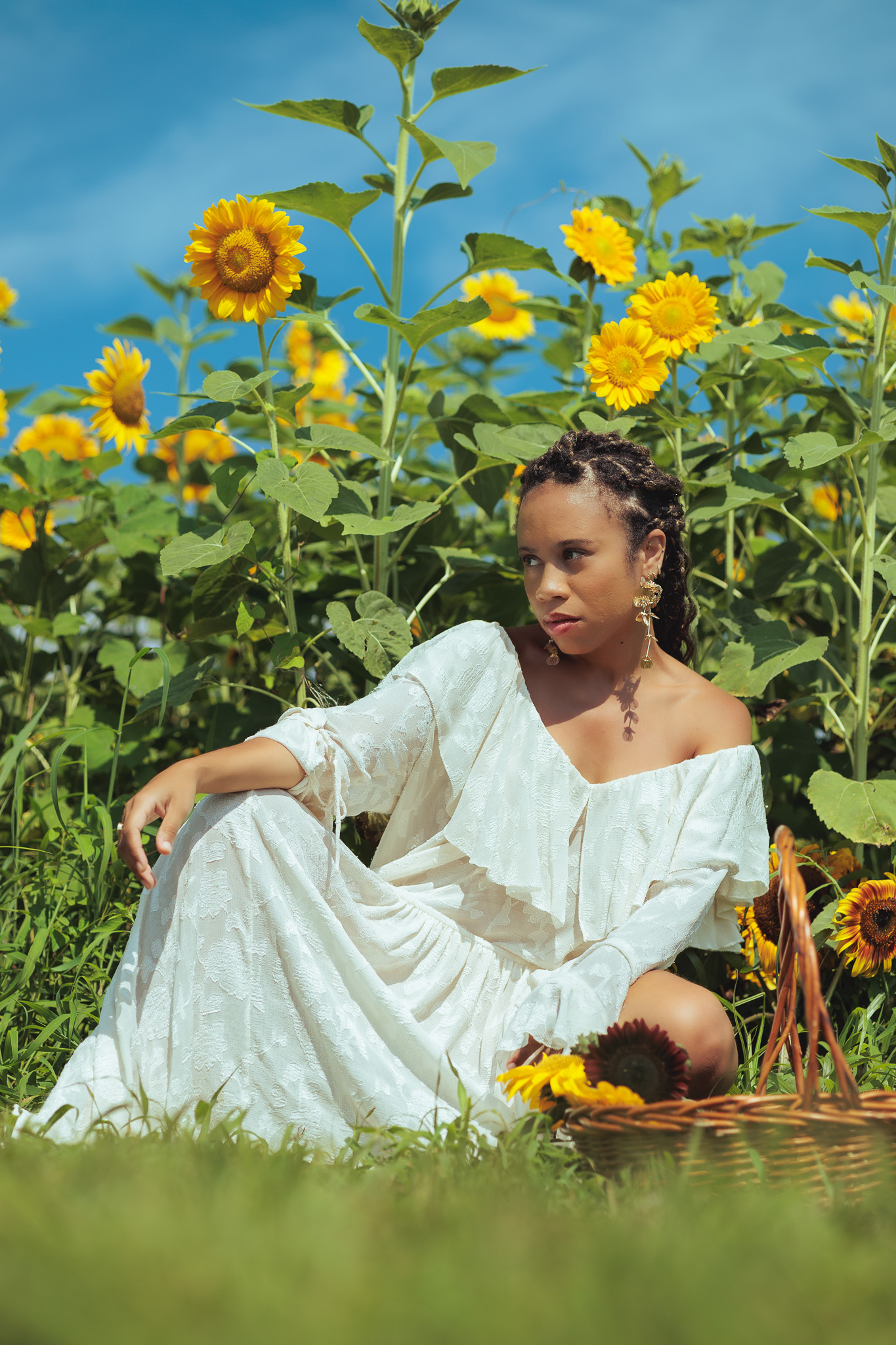 Woman in a white dress sitting in the sunflower field beside a basket of picked blooms